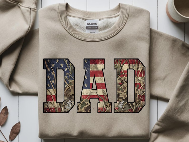 May include: A tan sweatshirt with the word "DAD" in large, block letters. The letters are filled with a distressed American flag design and camouflage pattern. The sweatshirt has a crew neck and long sleeves.