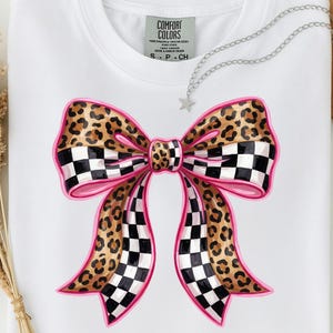 May include: White t-shirt with a large bow design. The bow features a leopard print pattern, pink outlines, and black and white checkered sections. The shirt has a Comfort Colors tag.