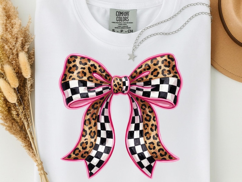 May include: White t-shirt with a large bow design. The bow features a leopard print pattern, pink outlines, and black and white checkered sections. The shirt has a Comfort Colors tag.