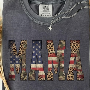 May include: Dark gray t-shirt with the word "MAMA" in large, bold letters. The letters are designed with a combination of American flag, camouflage, and leopard print patterns. A silver necklace with a star pendant is draped across the shirt.