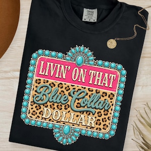 May include: Black t-shirt with a graphic design that reads "LIVIN' ON THAT Blue Collar DOLLAR." The design features a pink and leopard print background with turquoise accents. A gold necklace is draped on the shirt.