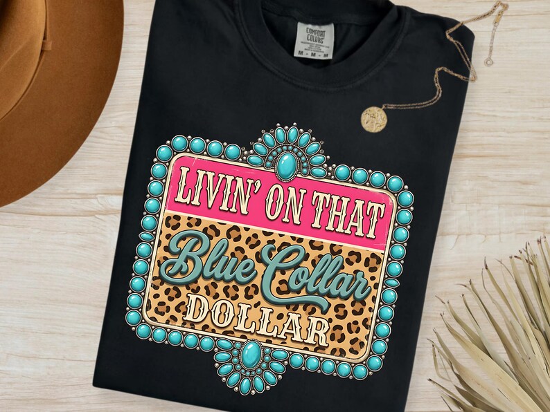 May include: Black t-shirt with a graphic design that reads "LIVIN' ON THAT Blue Collar DOLLAR." The design features a pink and leopard print background with turquoise accents. A gold necklace is draped on the shirt.