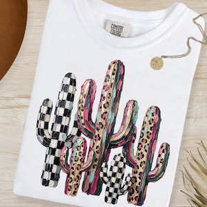 May include: White t-shirt featuring a colorful cactus design. The cactus illustration includes black and white checkered patterns, leopard print, and shades of pink, green, and brown. The shirt has a crew neck and a tag that reads "Comfort Colors".