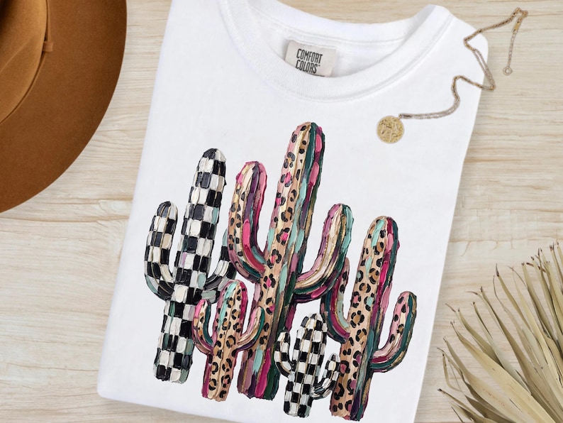 May include: White t-shirt featuring a colorful cactus design. The cactus illustration includes black and white checkered patterns, leopard print, and shades of pink, green, and brown. The shirt has a crew neck and a tag that reads "Comfort Colors".