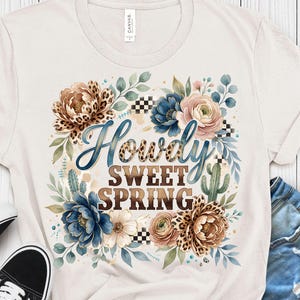May include: Cream-colored t-shirt with a floral design and the words "Howdy Sweet Spring." The design includes blue, pink, and brown flowers, green leaves, and a cactus. Black and white checkered pattern accents the design.