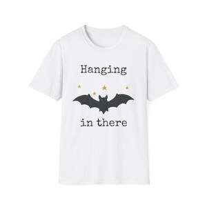 May include: A white short-sleeved t-shirt featuring a black graphic of a bat with outstretched wings. Above the bat, the text reads Hanging, and below the bat, the text reads in there. Four small gold-coloured stars are positioned above the bat.