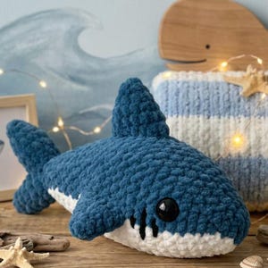 May include: A handmade blue and white crochet shark toy with black button eyes and a fin. The shark is lying on a wooden surface. The background includes a blue and white striped pillow, a wooden whale, and a painting of ocean waves.
