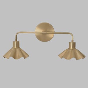 Pode incluir: Arandela de parede em tom de latão com duas luzes. A luminária possui uma placa traseira circular, uma barra horizontal e duas luzes voltadas para baixo com abajures em forma de sino e com babados. Design minimalista e elegante.