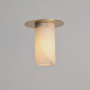 May include: A cylindrical pendant light fixture with a brass-colored top and a translucent, off-white shade. The light fixture is suspended from the ceiling, casting a soft glow. The design is modern and minimalist.