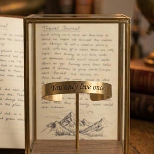 Puede incluir: Un brazalete dorado con la inscripción "You only live once" exhibido en una vitrina de vidrio y madera. La vitrina contiene un diario de viaje y una ilustración de montaña. Un reloj de bolsillo vintage y una etiqueta de cuero están sobre la superficie de madera.