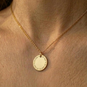 Puede incluir: Un collar dorado con una cadena delicada y un colgante circular. El colgante tiene texto grabado alrededor del borde. El collar se lleva alrededor del cuello.