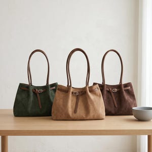 May include: Three suede tote bags in dark green, tan, and dark brown. Each bag features two long shoulder straps and a horizontal strap across the front secured with a silver-toned metal buckle. The bags have a soft, slouchy structure.