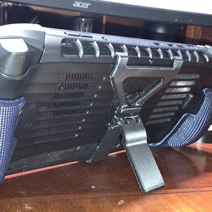 May include: A black and gray handheld gaming device with blue textured grips and a built-in stand. The device is resting on a wooden surface, with a computer monitor and other electronics in the background.