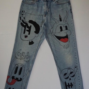 May include: Light blue denim jeans with unique cartoon illustrations in black. The designs include a character with a candle, a devil-like figure, a ghost, and a skull. The jeans have a classic five-pocket design.