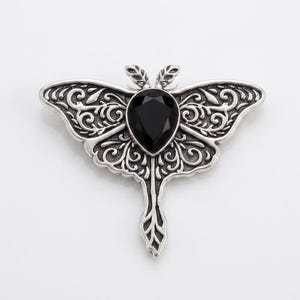 May include: A silver-toned moth brooch featuring intricate filigree scrollwork patterns on the wings and a central, faceted, pear-shaped black stone. The metal has a darkened finish in the recessed areas to highlight the detailed texture.