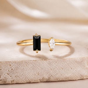 May include: A gold open ring featuring a rectangular black gemstone on one end and a marquise-cut clear gemstone on the other. The band is polished gold, and the two stones are positioned to face each other with a small gap between them.