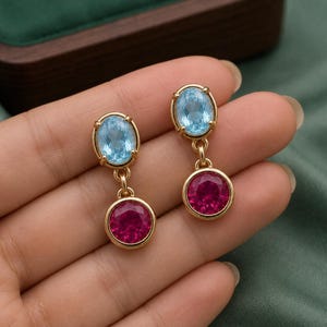 Peut inclure: Une paire de boucles d'oreilles pendantes en or avec une pierre précieuse ovale bleu clair à facettes en haut et une pierre ronde rose foncé à facettes suspendue en dessous. Chaque pierre est sertie dans une monture en or simple.