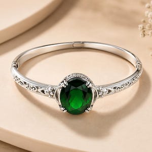 Może przedstawiać: Srebrna bransoletka typu bangle z zawiasem, ozdobiona owalnym, ciemnozielonym kamieniem szlachetnym pośrodku. Metalowa obręcz posiada po obu stronach skomplikowane filigranowe wzory oraz małe, przezroczyste kamienie osadzone przy oprawie.