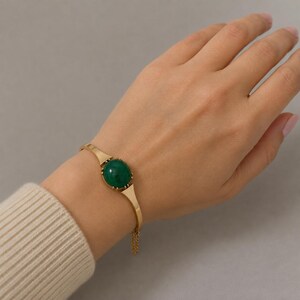 Op de afbeelding: Een goudkleurige metalen armband met een centrale, gladde, ronde, donkergroene cabochonsteen. De armband heeft een gepolijste afwerking en bevat een delicaat, bungelend goudkleurig kettinkje dat aan één kant van de metalen band is bevestigd.