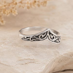 May include: A silver ring featuring a chevron or V-shaped silhouette with an oxidized finish. The band is decorated with intricate, raised scrollwork patterns that follow the pointed contour of the ring, creating a distinct contrast between the dark recessed areas and the polished silver surfaces.