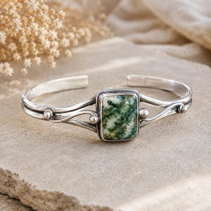 May include: A silver cuff bracelet featuring a rectangular moss agate stone with green and white inclusions. The metal band has a polished finish with decorative silver spheres and curved wire detailing on both sides of the central stone setting.