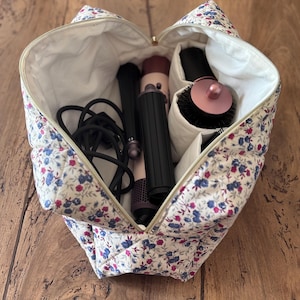 Op de afbeelding: Een gewatteerde cosmetische tas met een bloemenpatroon, open om haarstylingtools te laten zien. De tas is wit met blauwe en roze bloemen. Binnenin zitten een krultang, föhn en ronde borstel.