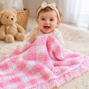 Può includere: Un bambino sorride mentre tiene in mano una coperta all'uncinetto rosa e bianca con frange. Un orsacchiotto e un cestino di vimini sono sullo sfondo. Il bambino indossa un completo color crema e una fascia con fiocco.