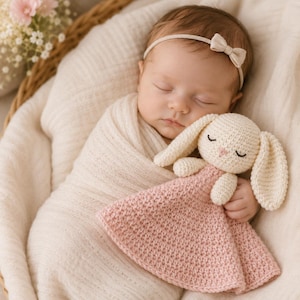 May include: A sleeping baby wrapped in a cream blanket, in a woven basket. The baby wears a bow headband and holds a crocheted bunny comforter with a pink body and cream head and ears. A soft, comforting nursery item.
