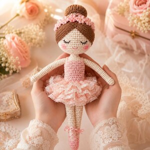 Puede incluir: Una muñeca de bailarina de ganchillo hecha a mano, con cabello castaño recogido en un moño y una diadema floral rosa. La muñeca lleva un maillot rosa y un tutú de encaje en capas. Zapatillas de ballet rosas con lazos completan el atuendo.