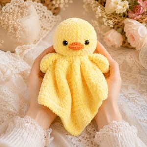 May include: A handmade yellow crochet duck toy with an orange beak and black eyes. The duck has a round head and a soft, textured body. The toy is designed to be a baby comfort item.