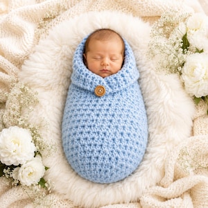 May include: A light blue crocheted baby swaddle sack featuring a textured star stitch pattern and a single wooden button closure at the top. The swaddle is designed to wrap around an infant, providing a snug fit while keeping the baby warm.
