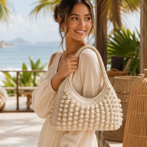 Può includere: Borsa a mano color crema, lavorata all'uncinetto, con un design a punto nocciola strutturato. La borsa ha una forma curva e un manico largo. Lo sfondo mostra una scena di spiaggia con palme e un vestito chiaro.