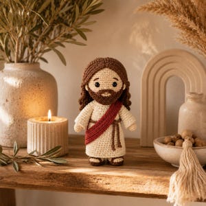May include: A crocheted figure of Jesus with brown hair and beard, wearing a cream-colored robe and a red sash. The handmade doll is standing on a wooden shelf, with a lit candle and decorative vases in the background.