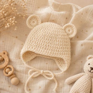 May include: Cream-colored, crocheted baby hat with bear ears and ties. The hat is made of soft yarn and has a textured appearance. A wooden toy and a stuffed bear are also in the image, suggesting a baby or nursery theme.