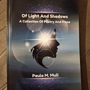 May include: A book titled "Of Light And Shadows" with the subtitle "A Collection Of Poetry And Prose." The cover features a silhouette of a face with a bright light emanating from behind it, set against a dark blue starry background. The author's name, Paula M. Mull, is at the bottom.