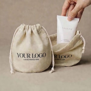 Op de afbeelding: Twee beige trekkoordzakjes, één gesloten en één open, met de tekst "YOUR LOGO CAN BE PRINTED HERE" in zwart gedrukt. De open zak bevat een witte tube "torana" brightening smoothing balm.