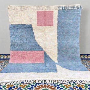 Puede incluir: Una alfombra rectangular con un patrón geométrico de bloques en color crema, rosa empolvado y azul claro. La alfombra tiene un pelo grueso y texturizado, y está rematada con flecos de color crema en los bordes superior e inferior.