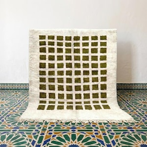 Puede incluir: Una alfombra rectangular con una base de color crema y un patrón de cuadrícula formado por cuadrados de color verde oliva oscuro. La alfombra tiene una textura de pelo largo y grueso, rematada con flecos cortos anudados en el borde inferior.