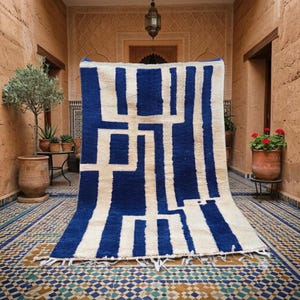 Puede incluir: Una alfombra rectangular con un patrón geométrico audaz de líneas verticales y horizontales de color azul intenso sobre un fondo crema. La alfombra tiene un pelo grueso y texturizado e incluye un fleco de borlas blancas en el borde inferior.