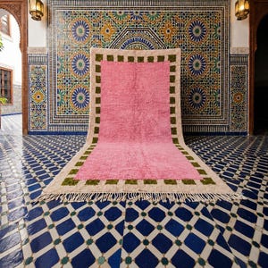 Puede incluir: Una alfombra rectangular que presenta un gran panel central de color rosa sólido rodeado por un borde ancho de color crema. El borde está acentuado con un patrón repetitivo de pequeños cuadrados verdes y acabado con flecos.