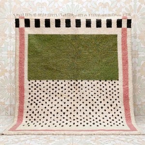 Puede incluir: Una alfombra rectangular con una base de color crema y un gran rectángulo verde oliva sólido en el centro. La alfombra está enmarcada por bordes verticales rosas y una banda horizontal superior con cuadrados alternos en negro y crema.