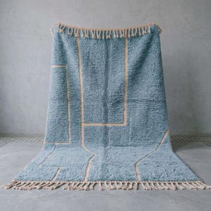 Puede incluir: Una alfombra rectangular con una textura de pelo largo en color azul y un patr&oacute;n geom&eacute;trico de l&iacute;neas finas en color beige claro. La alfombra presenta un fleco trenzado decorativo en los bordes superior e inferior del dise&ntilde;o.
