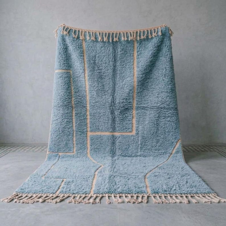 Puede incluir: Una alfombra rectangular con una textura de pelo largo en color azul y un patr&oacute;n geom&eacute;trico de l&iacute;neas finas en color beige claro. La alfombra presenta un fleco trenzado decorativo en los bordes superior e inferior del dise&ntilde;o.