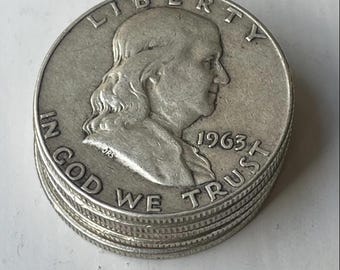 Pode incluir: Uma pilha de moedas de meio dólar Franklin de cor prateada. A moeda superior apresenta um retrato de perfil de Benjamin Franklin, o texto LIBERTY, a frase IN GOD WE TRUST e o ano 1963. As moedas têm uma borda serrilhada.