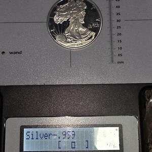May include: A silver coin with a detailed design of a woman walking towards the sun. The coin is on a gray surface with a ruler and a digital display showing "Silver-.999". The coin is being tested by a Precious Metal Verifier.