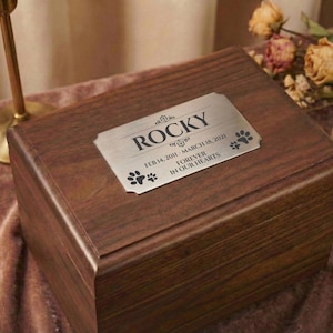 May include: A dark brown wooden pet urn with a silver nameplate. The nameplate reads "Rocky" with dates and the phrase "Forever in our hearts" and paw prints. The urn is rectangular and has a polished finish.