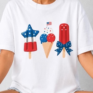 Puede incluir: Una camiseta blanca de manga corta con un estampado gráfico de tres dulces helados: un polo en forma de estrella con rayas rojas, blancas y azules, un cono de helado con bolas rojas y azules, y un polo rojo adornado con un lazo azul con estampado de estrellas.