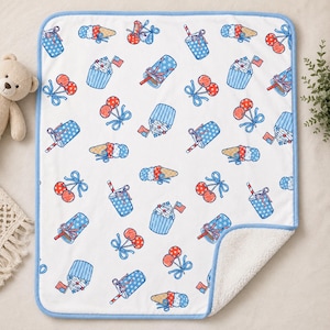 Puede incluir: Una manta rectangular blanca con borde azul claro y un patrón repetitivo de dulces patrióticos rojos, blancos y azules, incluyendo cupcakes con banderas, conos de helado, cerezas y bebidas con pajitas, con un reverso de sherpa blanco.