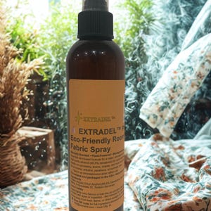 May include: A brown bottle of EXTRADEL Eco-Friendly Room Fabric Spray with a black spray nozzle. The label features text and a starburst design. The background includes greenery and patterned fabric.