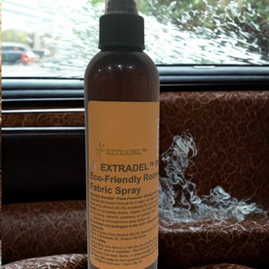 May include: A brown spray bottle with a black nozzle and a tan label. The label reads "EXTRADEL Eco-Friendly Room Fabric Spray". The bottle is in front of a car seat with a brown leather pattern.